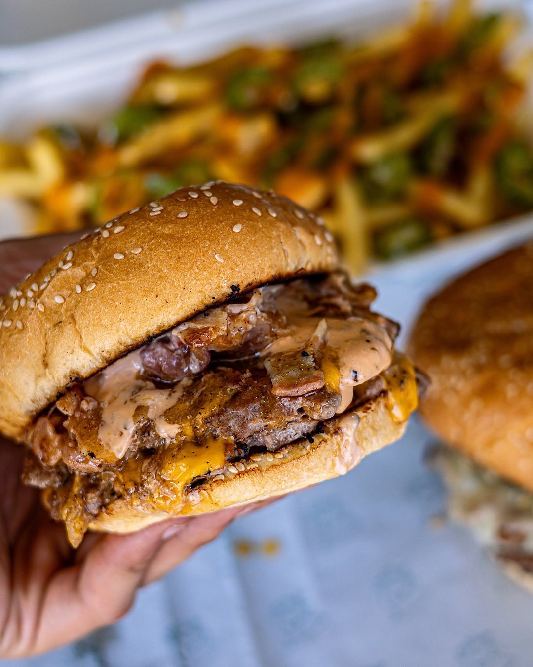A hand holding a Bulubeef double cheeseburger with bacon and sauce, loaded fries behind it